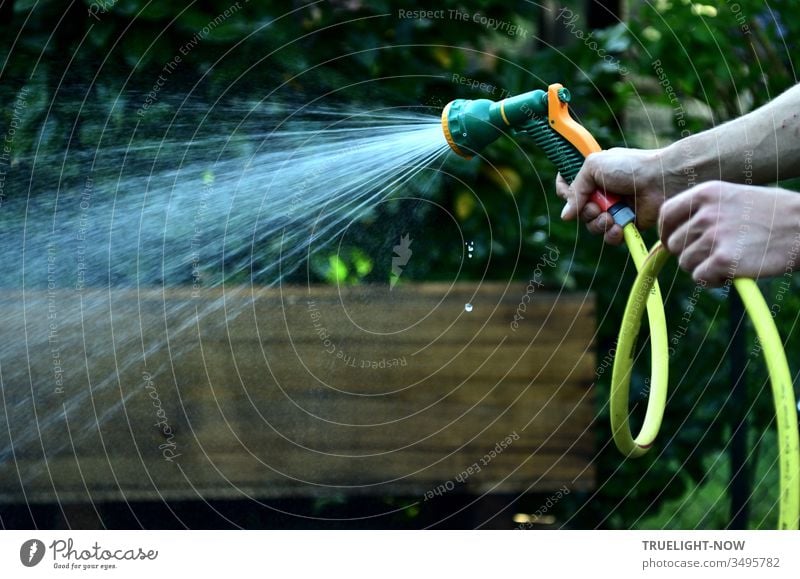 Zwei kräftige Hände vor braunem und dunkelgrünem Hintergrund halten einen gelben Gartenschlauch mit einem grün organge farbenen Duschkopf aus dem ein heller Brausestrahl frischen Wassers kommt, während einige kleine Tropfen senkrecht herabfallen