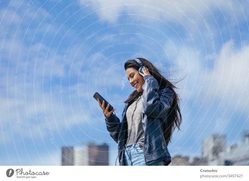 Braunes Mädchen lächelt und hört Musik mit einem blauen Helm, der mit dem Mobiltelefon verbunden ist Himmel Perspektive Lächeln genießen Tanzen urban Gebäude