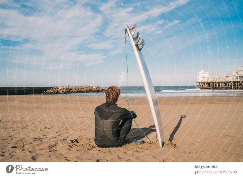 Surfer, der am Sandstrand und neben dem Surfbrett sitzt. Mann Brandung Wasser Sport Surfen MEER Meer im Freien sportlich Küstenlinie Wellen Hintergrund