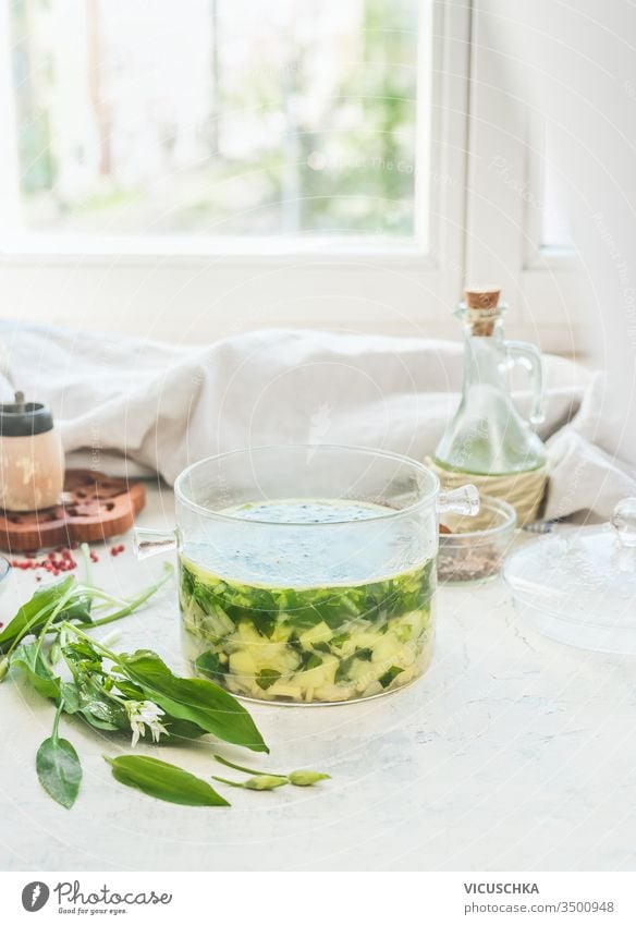 Grüne Bärlauchsuppe im Glaskochtopf auf hellem Tisch mit Zutaten im Fensterhintergrund stehen bleiben. Hausmannskost der Saison. Veganes Essen. Gesundes Kochen und Essen. Rezepte für den Frühling