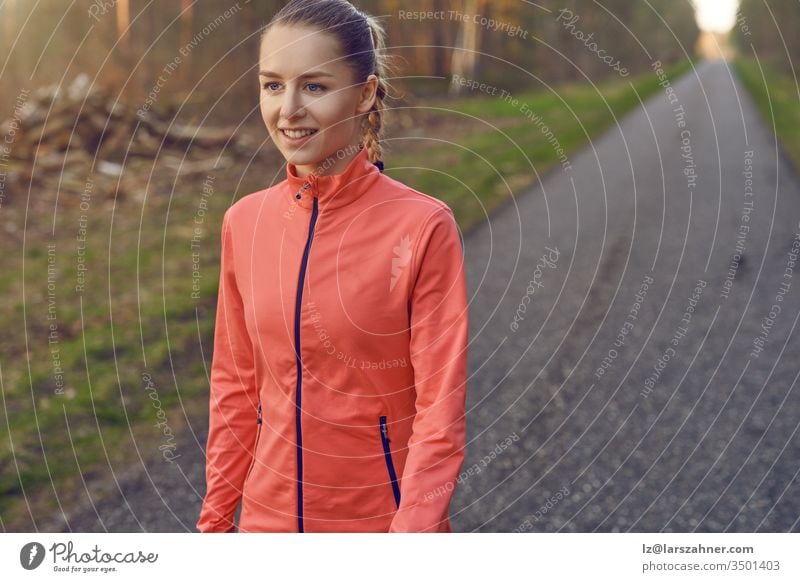 Lächelnde, sportlich-sportliche junge Frau, die auf einer geteerten Bahn durch Wälder, die vom warmen Schein der Sonne beleuchtet werden, in einem gesunden, aktiven Lebensstil trainiert