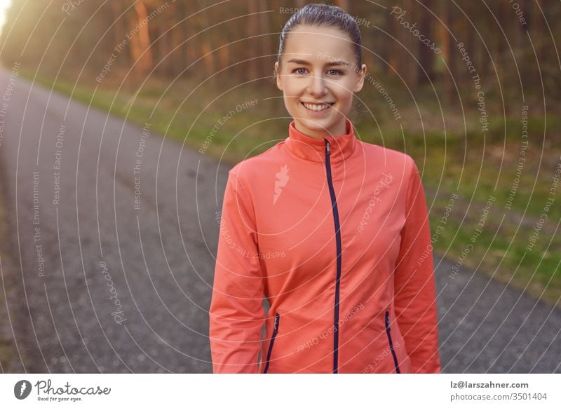 Lächelnde, sportlich-sportliche junge Frau, die auf einer geteerten Bahn durch Wälder, die vom warmen Schein der Sonne beleuchtet werden, in einem gesunden, aktiven Lebensstil trainiert