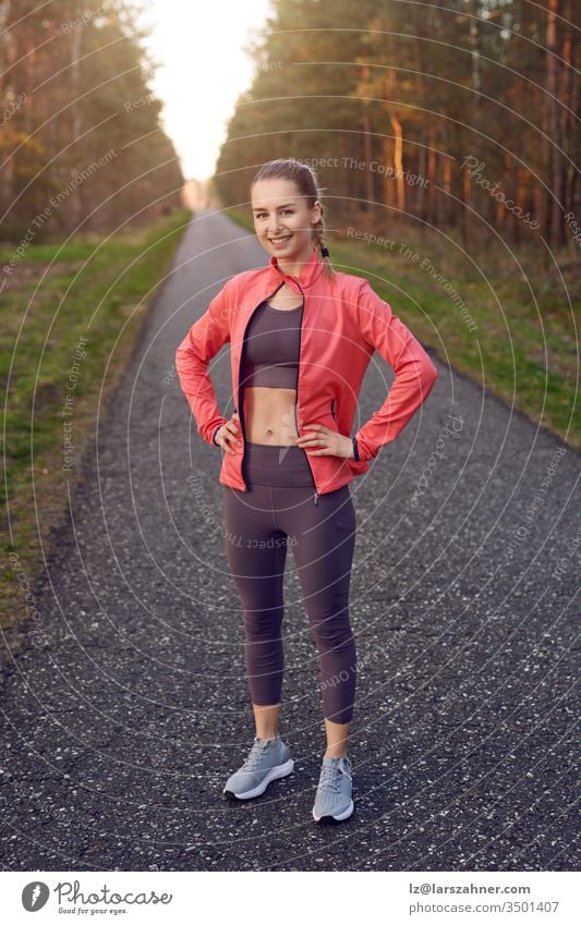 Ganzflächiges Frontporträt eines sportlichen jungen Mädchens in Trainingskleidung mit grauer Hose und roter Jacke, das nach dem Joggen mit den Händen auf den Hüften steht und sich ausruht, in die Kamera schaut und lächelt