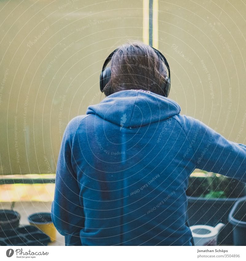 junge Frau sitzt mit Kopfhörern auf dem Balkon zum Hinterhof und pflanzt Blumen und Kräuter Ausblick Balconing Balkonbepflanzung Hobby Hoffnung Musik hören Saat