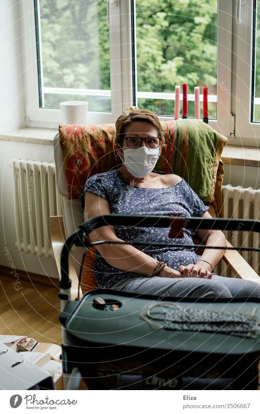 Frau der Risikogruppe mit Maske und Rollator sitzt aufgrund der Corona-Krise alleine zuhause in Isolation Covid-19 Mundschutz Blick in die Kamera isoliert