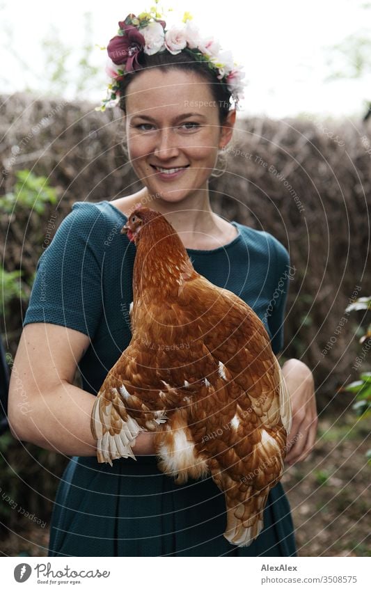 Junge Frau mit Blumenkranz im Haar steht im Hühnerauslauf hält braunes Huhn im Arm Zentralperspektive Schwache Tiefenschärfe Tag Außenaufnahme Farbfoto Idylle