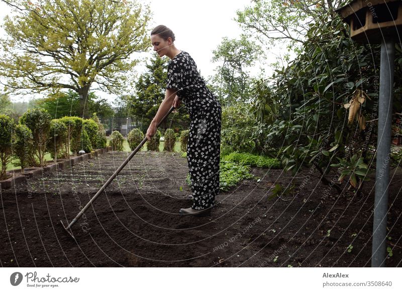 Junge Frau mit Blumenkranz im Haar harkt ein Gemüsebeet - Gartenarbeit Zentralperspektive Schwache Tiefenschärfe Tag Außenaufnahme Farbfoto Idylle feminin