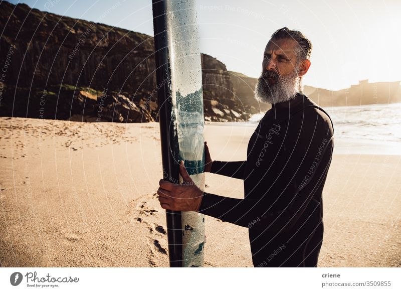 älterer Mann mit langem Bart trägt Surfbrett am Strand Senior Männer Urlaub Surfen Erwachsener Stehen graue Haare Lifestyle Beteiligung Sport Hobby Porträt