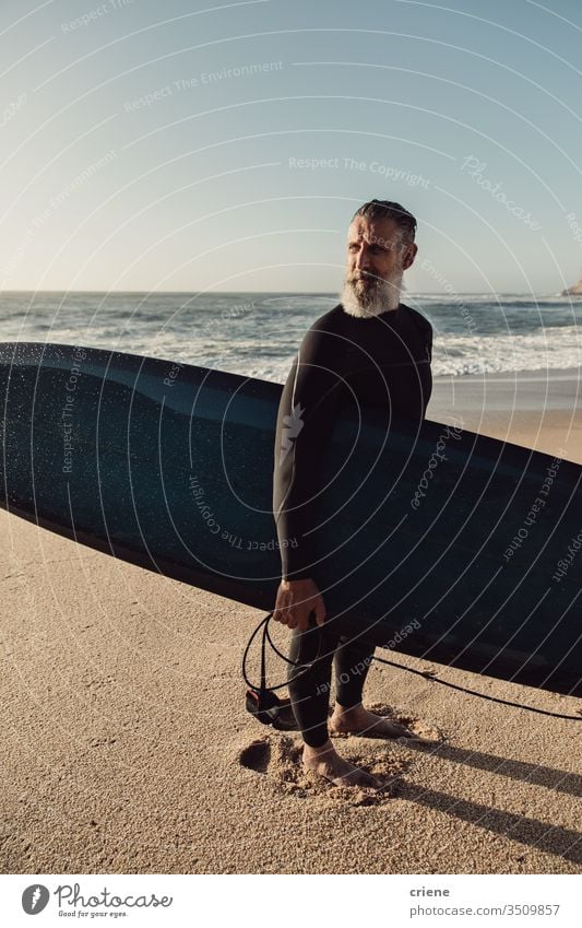 Lächelnder älterer Mann mit Surfbrett am Strand Senior Männer Urlaub Surfen Erwachsener graue Haare Lifestyle Freude Sport Hobby Porträt Vollbart Brandung Meer