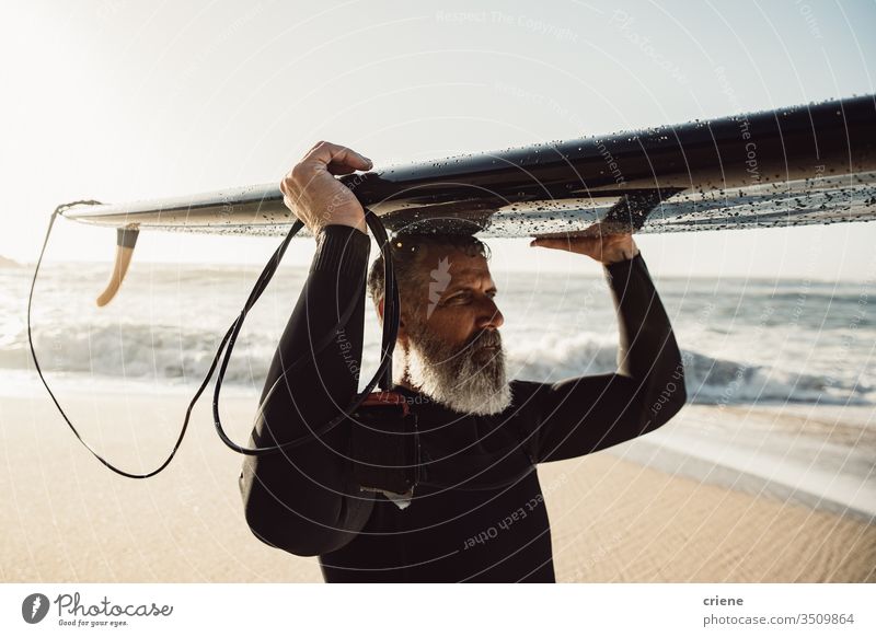 Kaukasischer älterer Mann mit Bart, der ein Surfbrett auf dem Kopf trägt Senior Männer Urlaub Strand Surfen Erwachsener graue Haare Lifestyle Sport Hobby