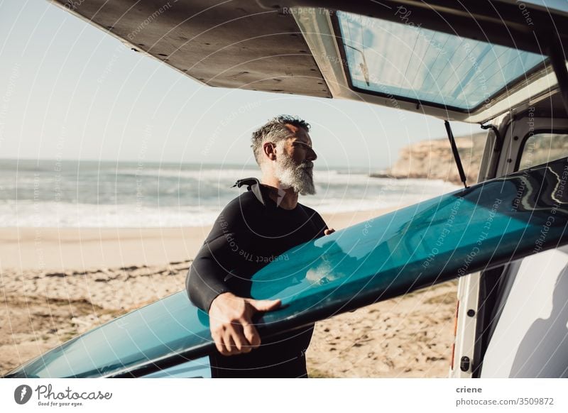 Älterer Mann nimmt am Strand Surfbrett aus Lieferwagen Senior Männer Kleintransporter Verkehr Ausflug Urlaub Surfen Erwachsener Vollbart graue Haare Lifestyle