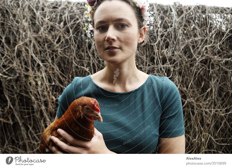 Junge Frau mit Blumenkranz im Haar steht im Hühnerauslauf hält braunes Huhn im Arm Zentralperspektive Schwache Tiefenschärfe Tag Außenaufnahme Farbfoto Idylle