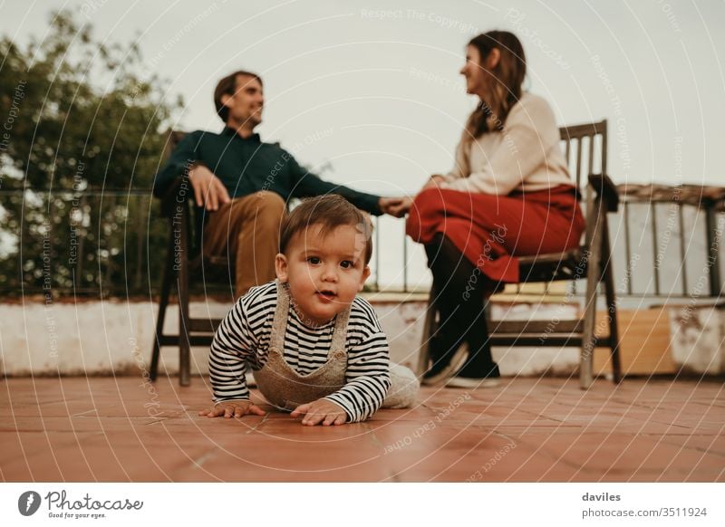 Süßer Säugling, der von seinen Eltern, die im Hintergrund sitzen, wegkrabbelt. Das Baby schaut in die Kamera. Vaterschaft niedlich Auge Haus heiter sprechend