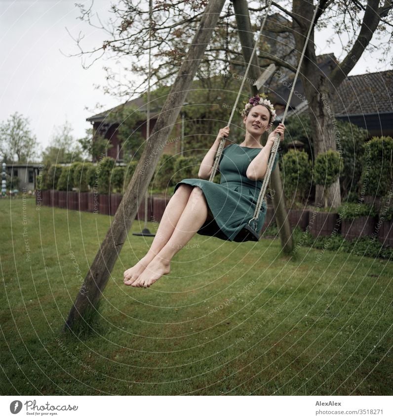 Analoges Portrait einer jungen Frau auf einer Schaukel im Garten mit Blumenkranz im Haar schön sportlich schlank grün Pflanze Hecke Stäucher Baum Natur