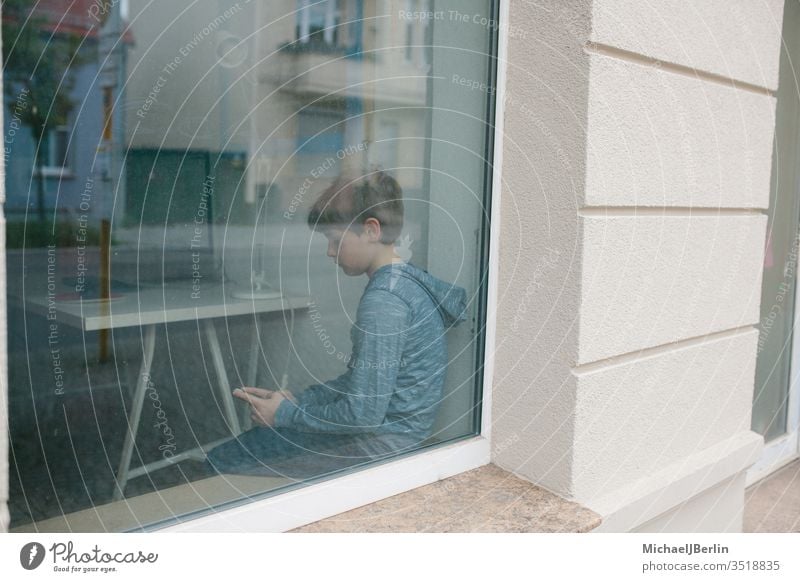 Junge sitzt einsam am Fenster und schaut auf sein Handy während Lockdown Isolation in der Coronavirus Epidemie Kind einsamkeit alleine fenster fensterbank