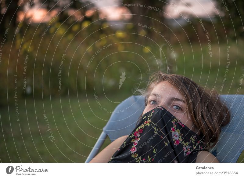 Abendstimmung im Garten, die brünette,  junge Frau im Liegestuhl hat ihr T-shirt über Mund und Nase gezogen und schaut in die Kamera feminin Gesicht Porträt