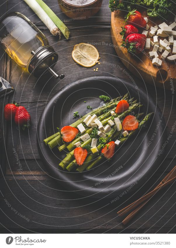 Schmackhafter Spargelsalat, belegt mit Erdbeeren und Frischkäsewürfeln, serviert in schwarzer Platte auf dunklem, rustikalem Tischhintergrund mit Zutaten. Ansicht von oben. Gesundes Essen. Hausmannskost und Essen