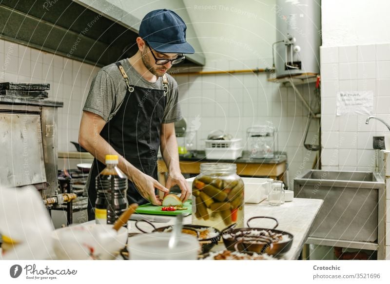 Mann mit Brille und Mütze kocht in einer Restaurantküche mit Töpfen mit Essen, Öl und Pfannen wirtschaftlich Container Urelemente professionell rostfrei Geräte