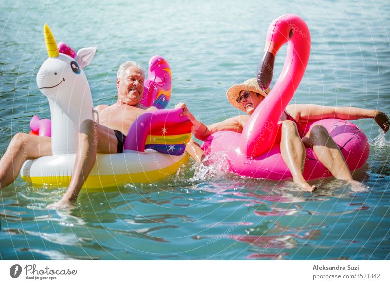 Älteres Paar amüsiert sich auf aufblasbaren Flamingos und Einhörnern. Lustige aktive Rentner, die zusammen glücklich den Sommerurlaub am Strand in Europa genießen, lachen, den Narren spielen, Wasser spritzen.