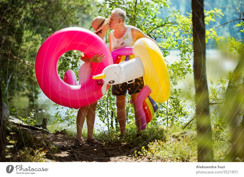 Ein älteres Ehepaar, das auf einem Waldweg am Meeresufer spazieren geht und einen aufblasbaren Riesenflamingo und ein Einhorn in der Hand hält. Lustige aktive Rentner genießen Sommerurlaub am Strand in Nordeuropa