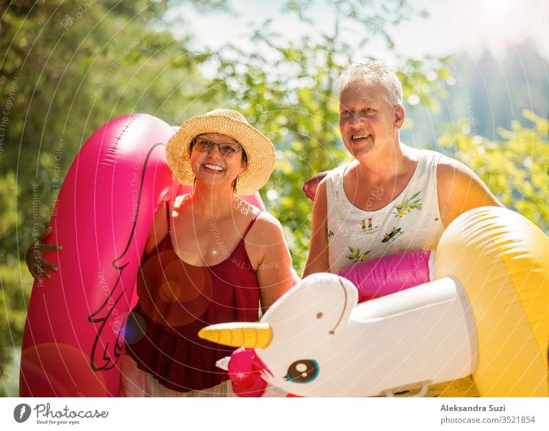 Ein älteres Ehepaar, das auf einem Waldweg am Meeresufer spazieren geht und einen aufblasbaren Riesenflamingo und ein Einhorn in der Hand hält. Lustige aktive Rentner genießen Sommerurlaub am Strand in Nordeuropa