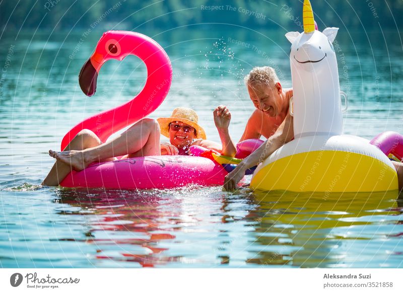 Älteres Paar amüsiert sich auf aufblasbaren Flamingos und Einhörnern. Lustige aktive Rentner, die zusammen glücklich den Sommerurlaub am Strand in Europa genießen, lachen, den Narren spielen, Wasser spritzen.