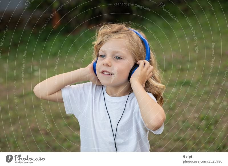 Lustiges Kind mit langen Haaren, das mit blauen Hadphones Musik hört Park hören Kindheit freudig Kopfhörer Spaß Behaarung blond Gesundheit Person Fröhlichkeit