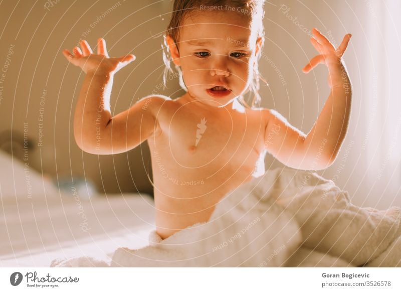 Süßes gelocktes kleines Mädchen mit einem Handtuch über dem nassen Körper, das nach dem Duschen in einem Schlafzimmer sitzt bezaubernd Baby Bad Bett Pflege