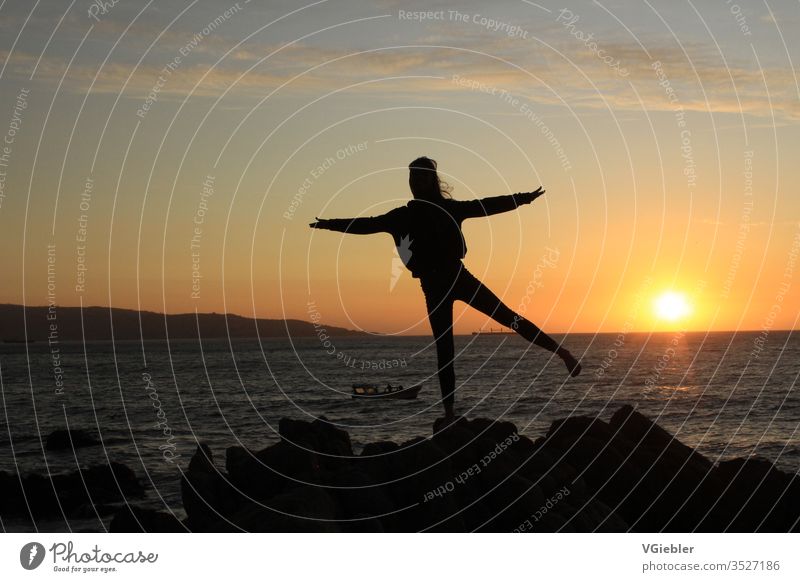 Umriss von Mädchen vor Sonnenuntergang, steht auf felsigem Untergrund, im Hintergrund das Meer und ein Boot Küste Sihouette Glück Freude Himmel Sommer Aktion