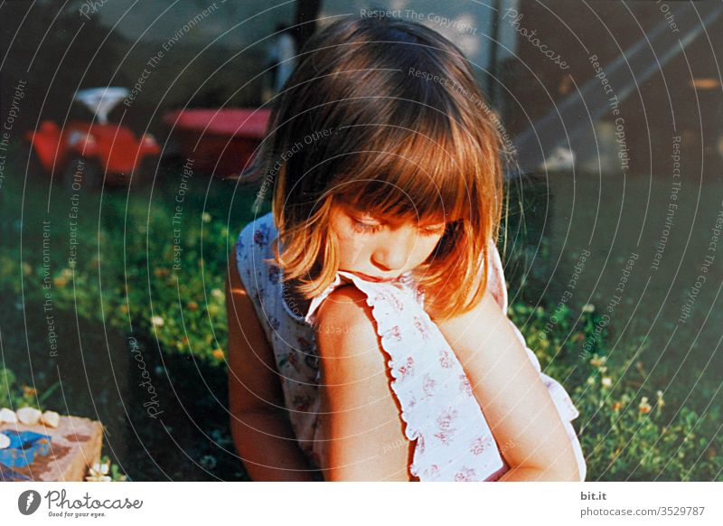 Nachdenkliches Mädchen im Kleid, sitzt auf der Wiese im Garten mit Spielzeug im Hintergrund und träumt, von Sonnenlicht bestrahlt Kind Kindheit Bobbycar Sommer