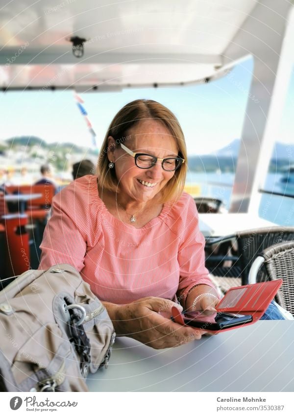 Ü70 Frau überprüft freudige Nachrichten auf dem Smartphone während einer Schifffahrt Seniorin pensioniert pensionärin bootsfahrt lachen Modus modisch verrotten