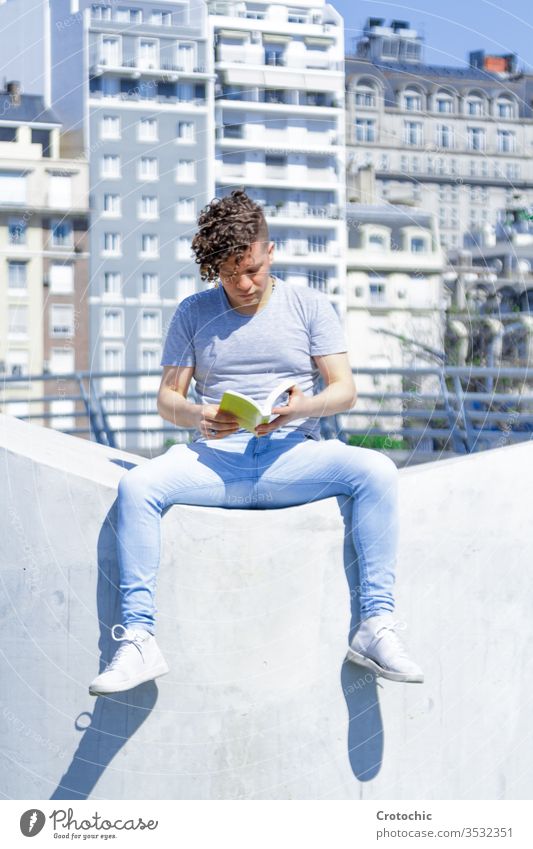 Mann mit moderner Frisur sitzt auf einer weißen Wand und liest ein Buch vertikal Großstadt urban Sitzen Park Straße sonnig Sonne Sonnenbad Sommer