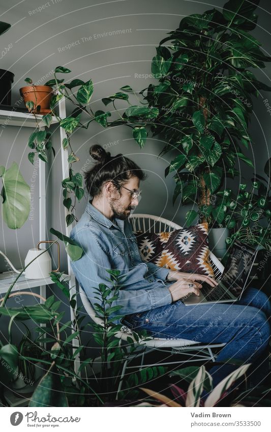 Junger Mann mit Bart sitzt auf dem Stuhl und benutzt einen Laptop. Freiberufliche Arbeit von zu Hause im Quarantänekonzept Appartement attraktiv Browsen