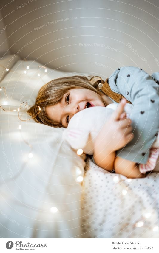 Glückliches Mädchen spielt auf weichem Bett spielen Spielzeug Umarmung Lachen Lichterkette Plüsch Lügen aufgeregt Kind Liebe Girlande Schlafzimmer wenig