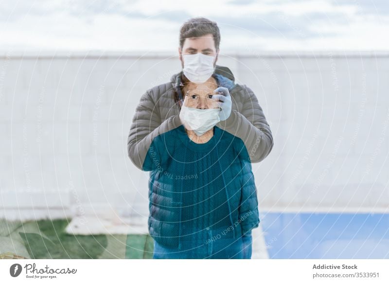 Junger Typ mit medizinischer Maske fotografiert ältere Frau mit medizinischer Maske durch Glas Großmutter Quarantäne Selbstisolierung separat fotografieren