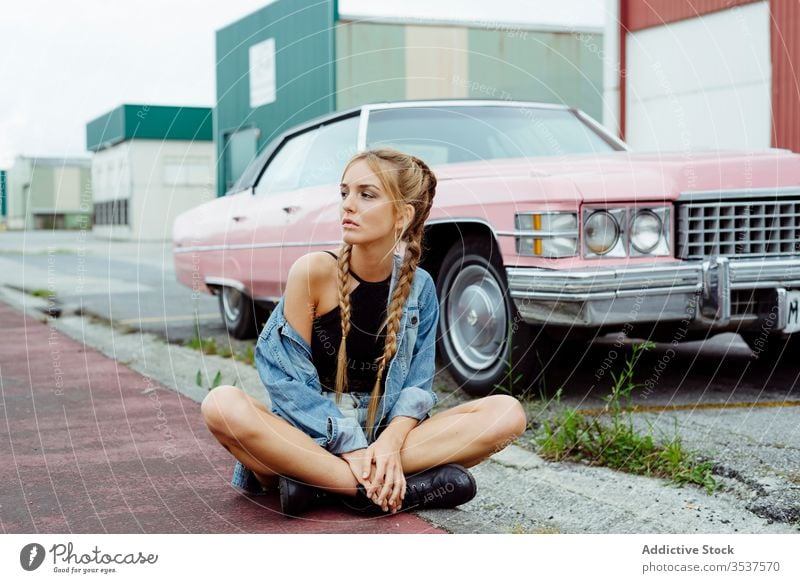 Attraktives blondes Mädchen sitzt auf dem Bürgersteig in der Nähe eines rosa Oldtimers Frau jung Zopf Sitzen PKW klassisch alt Grunge Sommer Porträt Freizeit