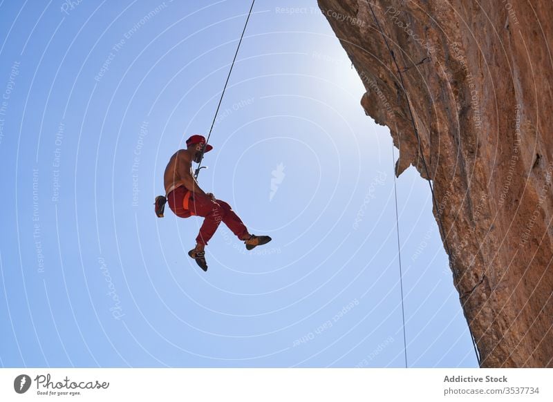 Starker männlicher Kletterer hängt an einem Seil in der Nähe eines felsigen Berghangs an einem sonnigen Tag Mann Bergsteiger stark Sport hängen sicheres Gerät