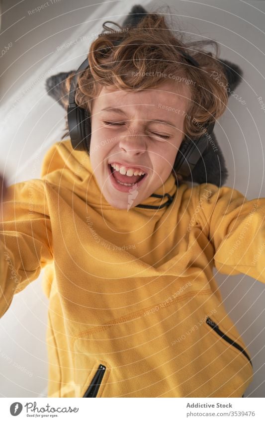 Zufriedenes Kind mit Kopfhörern auf dem Tisch liegend und Musik in hellem, modernem Raum genießend Junge Kälte zuhören heimwärts Glück Gesang Freude