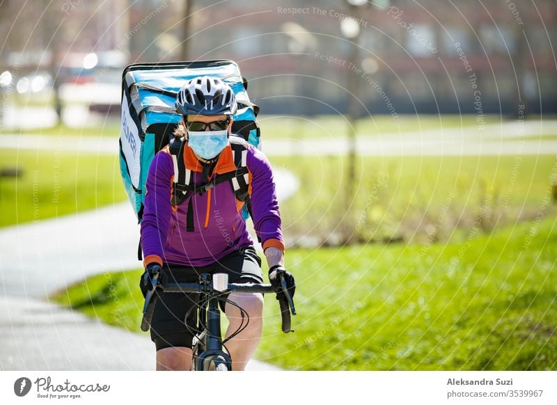 Frau mit Helm und Schutzmaske mit großem Essenslieferungsrucksack mit Fahrrad auf der Straße. Sonniger Frühlingstag in der Stadt. Porträt eines weiblichen Kuriers in einer medizinischen Maske.