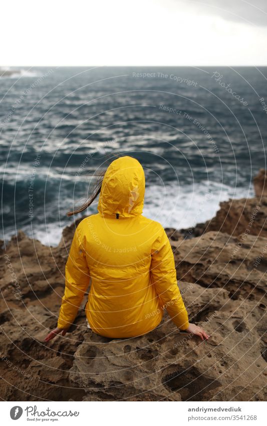 junge Frau im gelben Regenmantel auf der Klippe sitzend und auf die großen Wellen des Meeres schauend, während sie an einem Regentag am Felsstrand bei bewölktem Frühlingswetter die schöne Meereslandschaft genießt