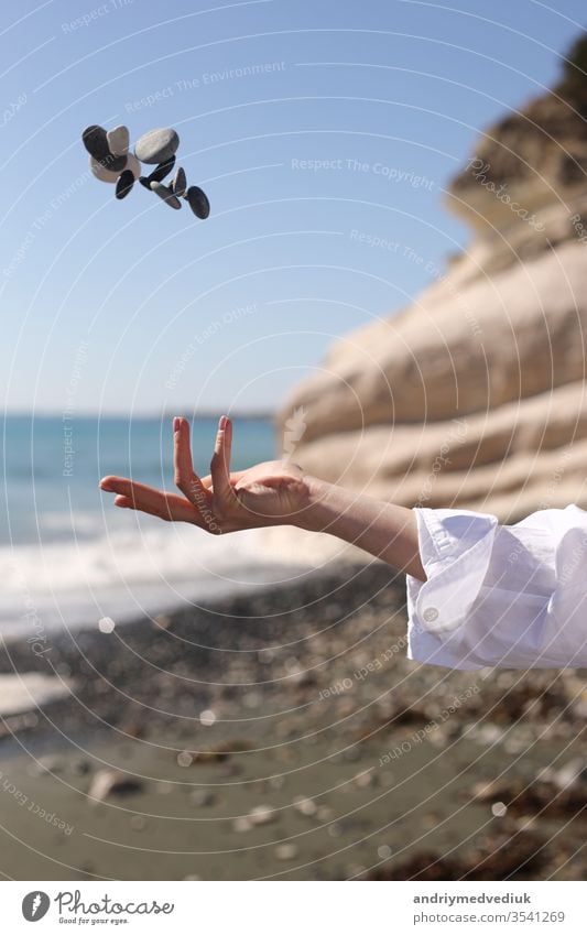 weibliche Hand wirft einen Kieselstein in die Hand vor dem Hintergrund des blauen Himmels, des Strandes, der Felsen und des Meeres. Frau MEER Mädchen