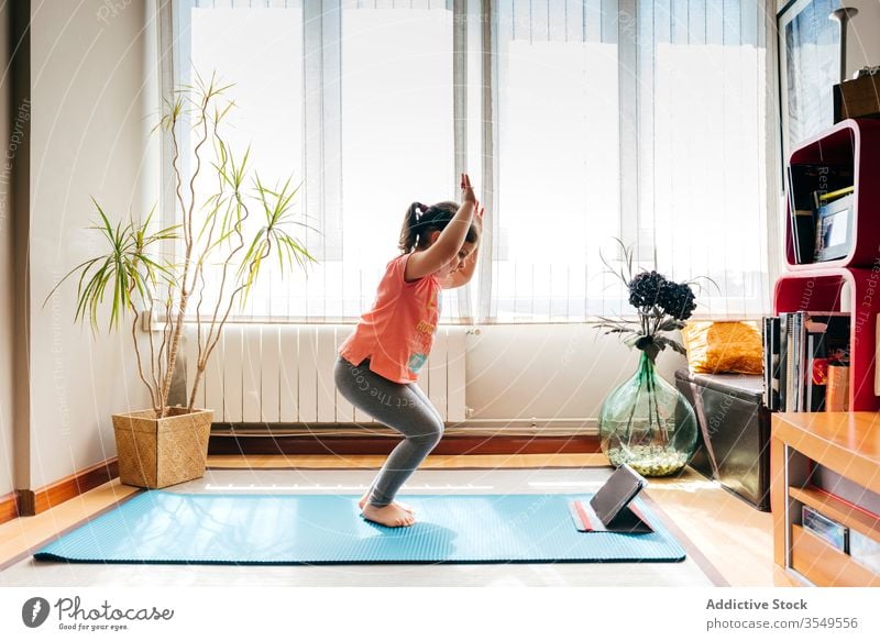 Fröhliches kleines Mädchen macht Yoga im hellen Raum Kind Pose Unterlage Video Tablette Tutorial Gleichgewicht üben zu Hause Stressabbau zuschauen Harmonie