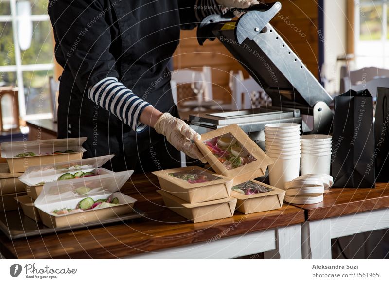 Restaurantmitarbeiter mit Schutzmaske und Handschuhen, die in Lebensmittelkartons verpacktes Essen zum Mitnehmen verpacken. Lebensmittellieferdienste und kontaktloser Online-Einkauf von Lebensmitteln.