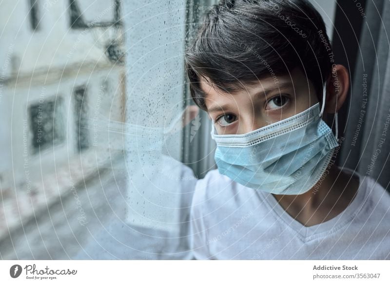 Trauriger Junge mit medizinischer Maske am Fenster Mundschutz Coronavirus Seuche heimwärts traurig unglücklich Selbstisolierung soziale Distanzierung Kind