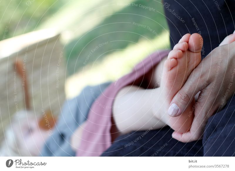 Hand der Mutter hält zärtlich, fürsorglich, liebevoll den nackten Fuß, des schlafenden Kindes mit Schnuller, während des Einschlafen, draussen am Schlafplatz in der Natur. Das Kleinkind schläft auf einer Wiese im Garten und träumt friedlich.