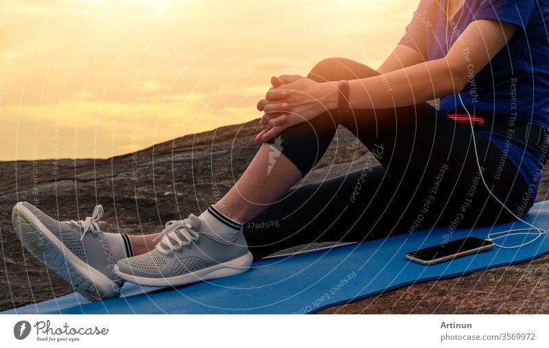 Frau sitzt und entspannt auf Yogamatte und hört Musik über Smartphone am Steinstrand am Meer. Frau trainiert im Freien. Fites Mädchen trägt Smartband. Gesunder, entspannter Lebensstil. Entspannen in den Sommerferien.