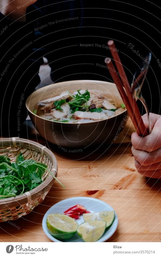 Eine Person sitzt mit Essstäbchen in der Hand vor einer leckeren Pho Suppe in Vietnam Essen Stäbchen Limette frisch heiß Kräuter traditionell Mahlzeit Schüssel