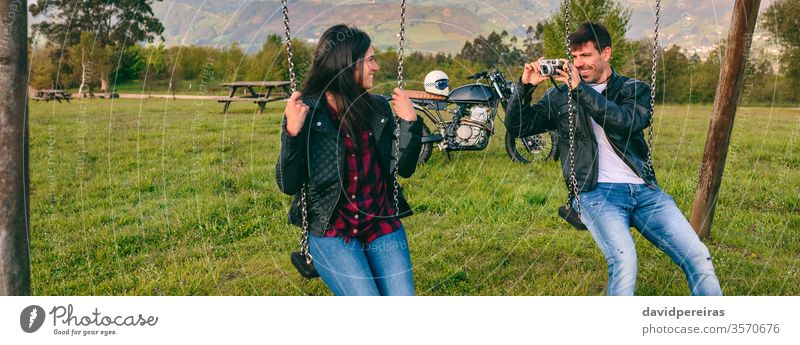 Junger Mann macht ein Foto von seiner Freundin auf der Schaukel Paar fotografierend Fotokamera pendeln Glück Motorrad Lederjacke Biker Transparente Netz