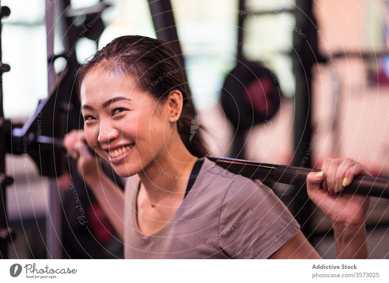 Kräftige junge asiatische Sportlerin macht Kniebeuge mit Langhantel in moderner Turnhalle Frau Curl-Hantel Training aktive Kleidung Maschine Gerät Fokus atmen