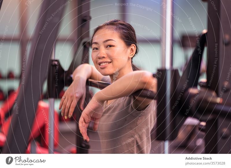 Positive asiatische Sportlerin lehnt sich in moderner Turnhalle an Langhantel und schaut in die Kamera Frau Curl-Hantel ruhen Training Fitnessstudio Maschine